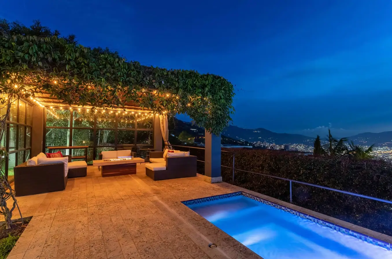 Outdoor seating area on a terrace with string lights, overlooking a city at dusk and a rectangular illuminated pool beside it.
